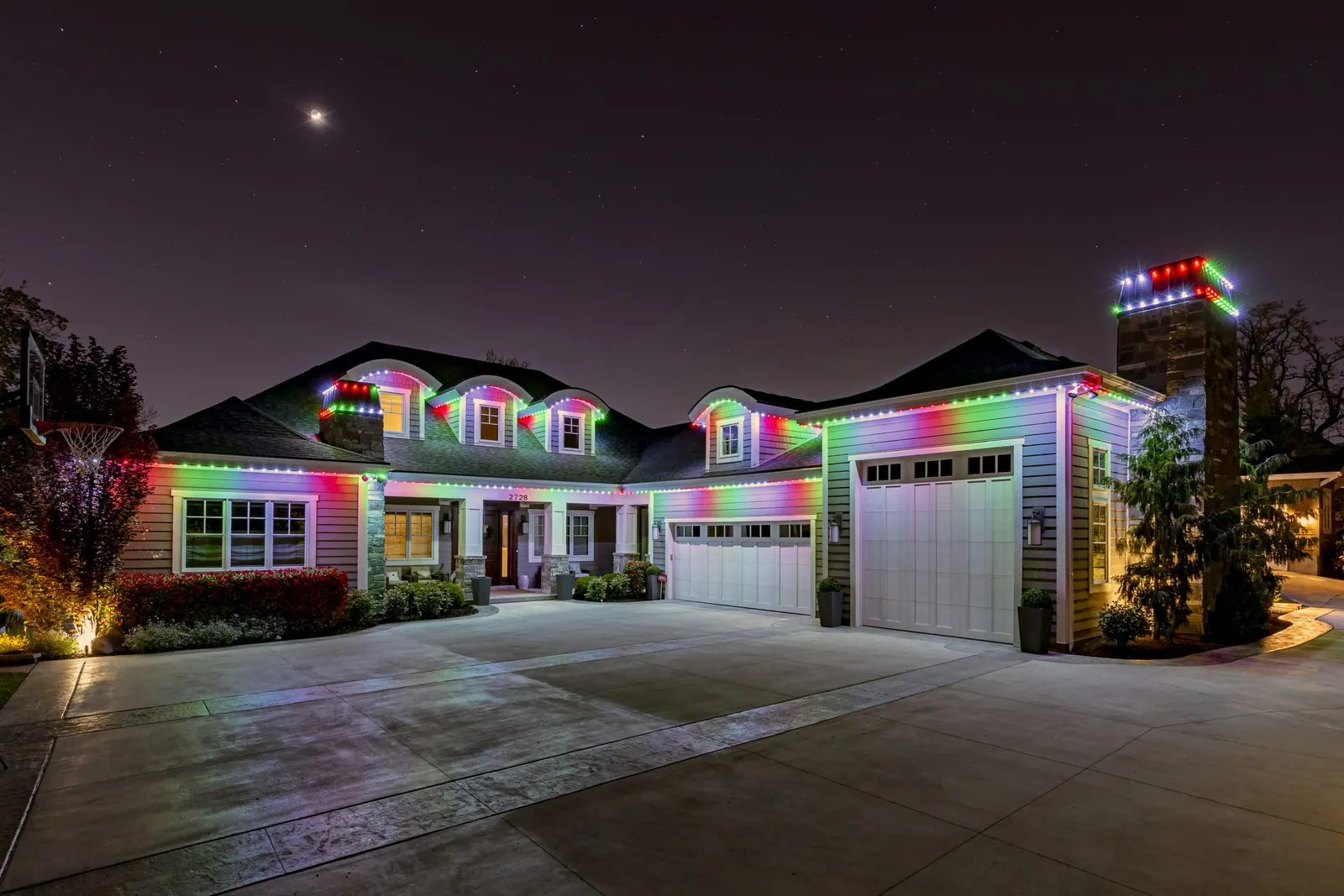 Large two-storey home with dormers washed in red, green and white permanent LED holiday lighting under a starry sky