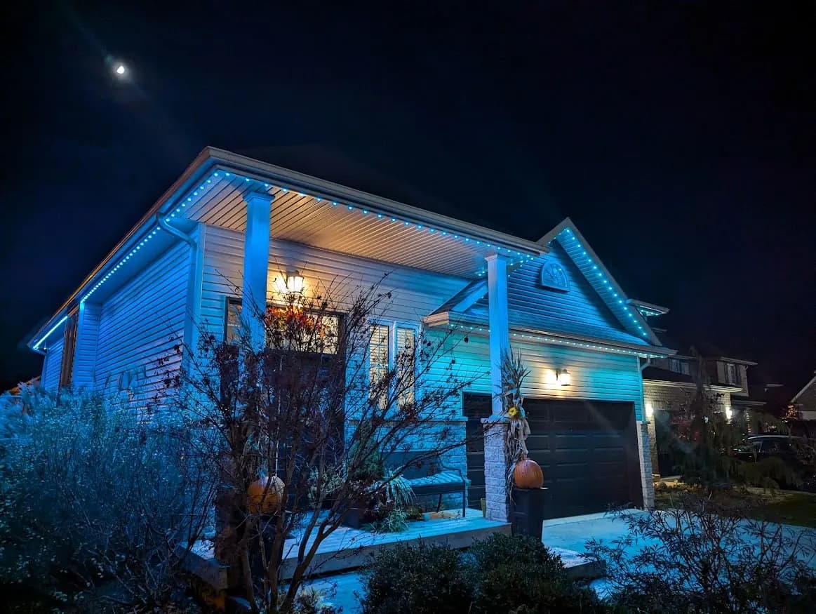 Bungalow with white siding lit in blue by permanent LED soffit lights at night, pumpkins on the porch - permanent lighting installation in London, Ontario