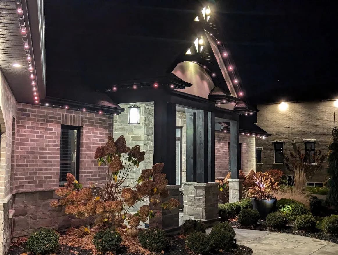 Stone and brick home with a steep black gable outlined in soft pink permanent LED lights at night - permanent roofline lighting in London, Ontario