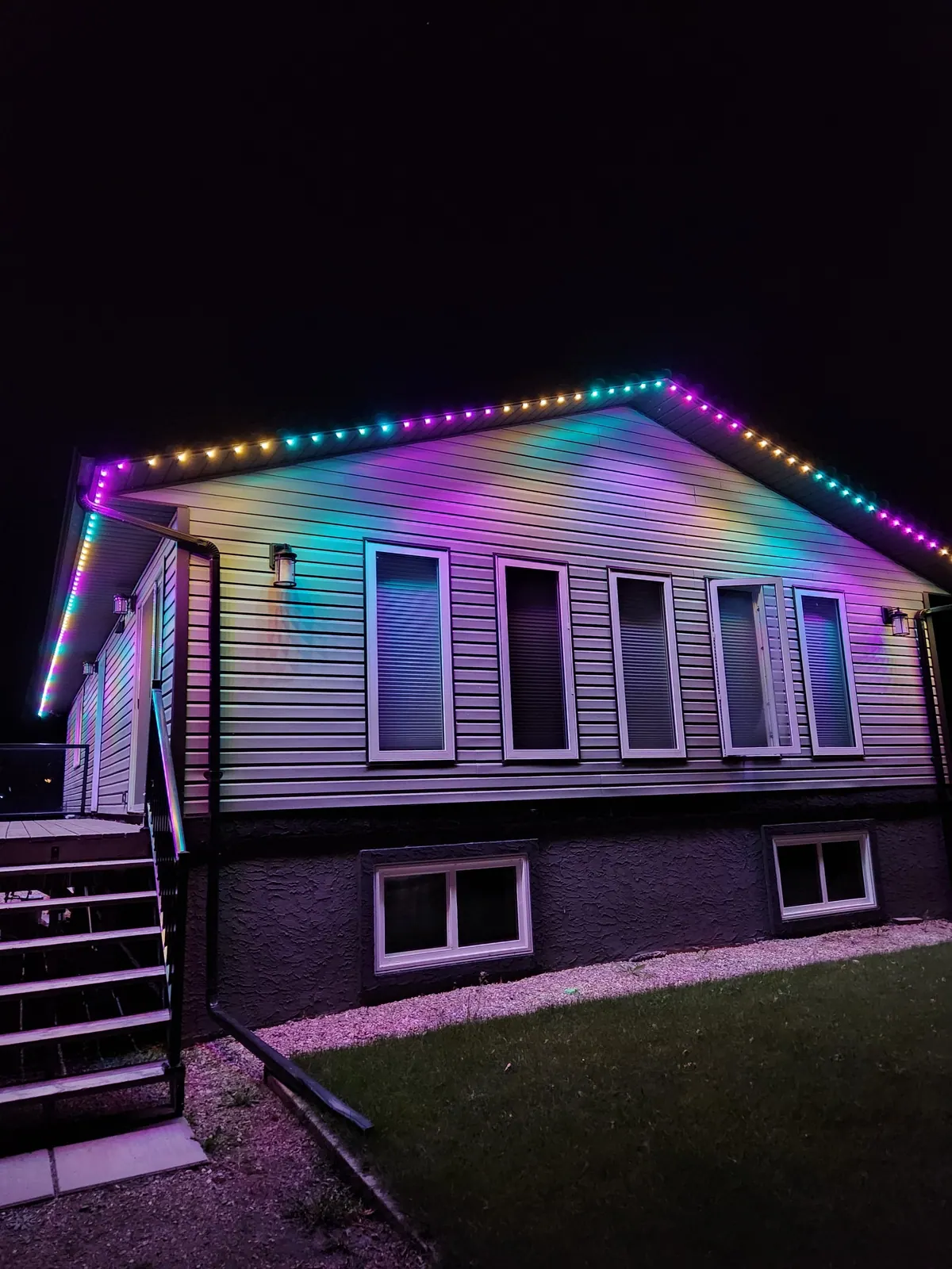 Bungalow with vinyl siding washed in a flowing rainbow gradient from permanent LED roofline lights at night