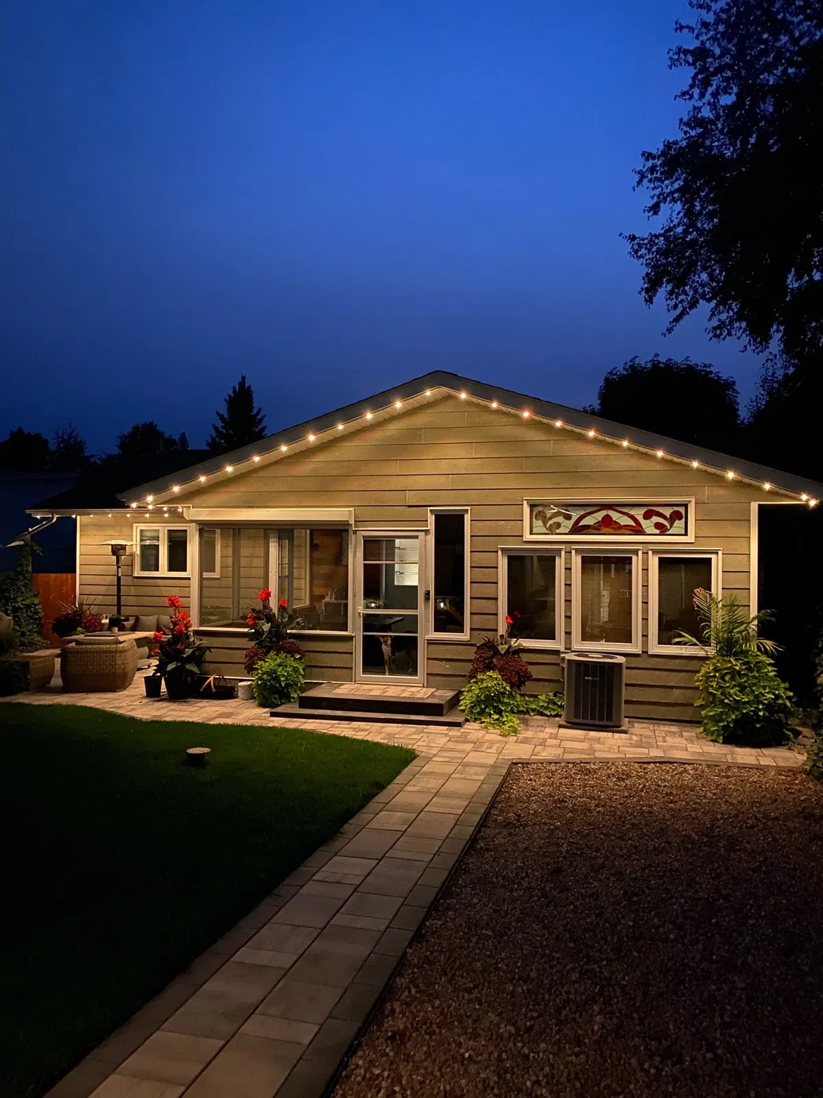 Backyard view of a bungalow at dusk with warm white permanent LED lights along the roofline above a patio and garden