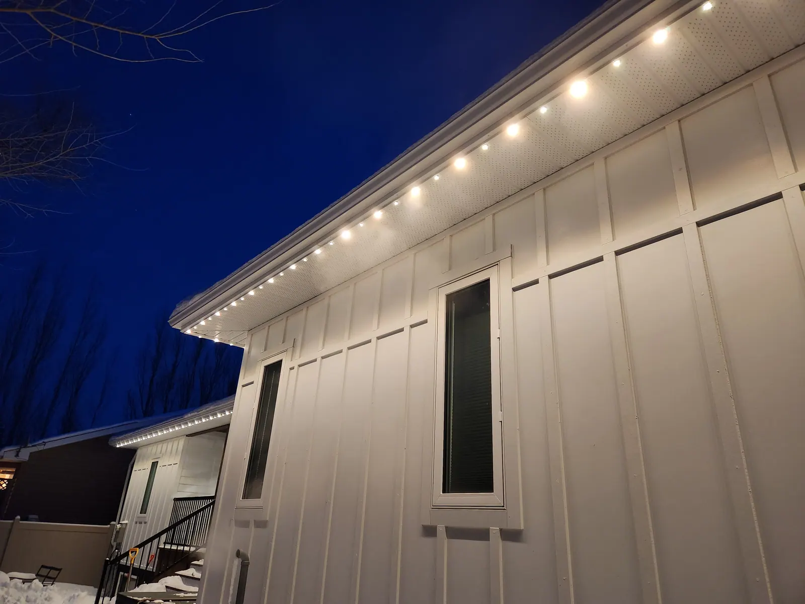 Close-up of warm white permanent LED soffit lights glowing under a snow-covered eave on a white board-and-batten home